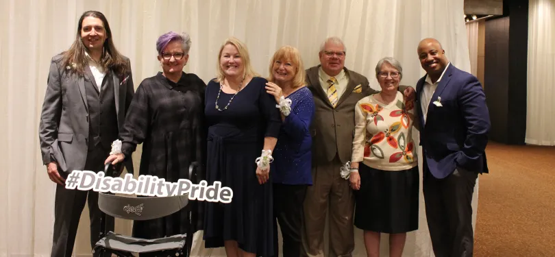 Seven community heroes standing in a line with a Disability Pride sign in front of the person standing 2nd from the left.